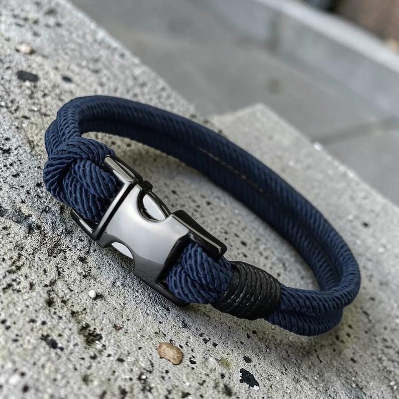 Puede incluir: Pulsera de cuerda azul marino con cierre met&aacute;lico plateado. La pulsera est&aacute; hecha de un material de cuerda gruesa y tejida. El cierre tiene forma rectangular con un cierre seguro. La pulsera se muestra sobre una superficie de hormig&oacute;n gris.