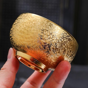 May include: A gold-colored bowl with a textured surface. The bowl has a rounded shape and a small, flat base. The bowl is held in a hand, with a dark background.