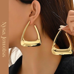 Puede incluir: Pendientes geométricos dorados con diseño triangular. Los pendientes tienen una superficie pulida y reflectante. También se incluye el texto "Nysa Jewelss".