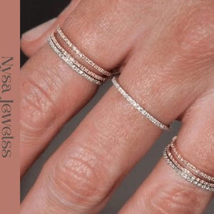 May include: Close-up of several diamond rings on a hand. The rings are delicate, with small, sparkling stones set in a thin band. One ring is a single band, while others feature multiple bands. The rings are set against a skin-toned background.