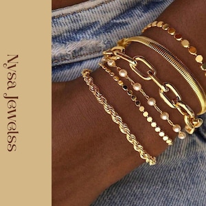 Puede incluir: Una colección de pulseras doradas de varios estilos, incluyendo una cadena de cuerda, una cadena con eslabones grandes y una pulsera con pequeños detalles redondos. Las pulseras están apiladas en una muñeca, con el texto "Nysa Jewelss" visible.