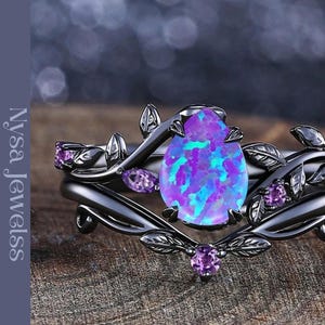 May include: A black ring set with a large, oval, purple and blue opal center stone. The ring is adorned with leaf and vine details and smaller purple accent stones. The brand name "Nysa Jewelss" is printed on the left side.