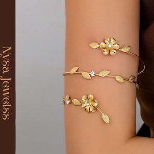 May include: Gold-toned arm cuff jewelry featuring floral and leaf designs. The cuff wraps around the arm with delicate leaves and sparkling accents. The jewelry is displayed on an arm, showcasing the intricate details and craftsmanship.