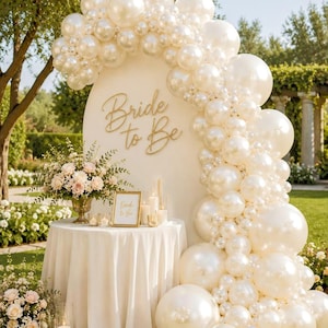 May include: A cream-colored arched backdrop featuring the gold script text Bride to Be, adorned with a large balloon garland of glossy white and pearl-finish balloons. A round table with a white tablecloth holds a floral arrangement and a sign.