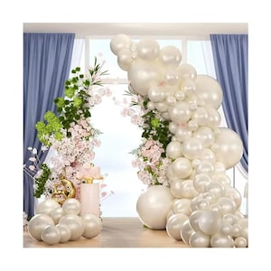 May include: A decorative arrangement featuring a white balloon arch, complemented by floral accents and a small cake. The scene is set against a window with blue curtains, creating an elegant and festive atmosphere, perfect for celebrations.