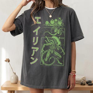 May include: Dark gray t-shirt featuring a green and black graphic of an alien creature with the Japanese word for "Alien". The design includes alien eggs and a detailed illustration of the alien. The shirt has a vintage, worn look.