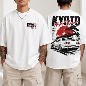 May include: White t-shirt with a graphic design featuring the word "KYOTO" and Japanese text. The back of the shirt displays a red sun, a car, and the words "KYOTO Night Runner" in black. The shirt has short sleeves.