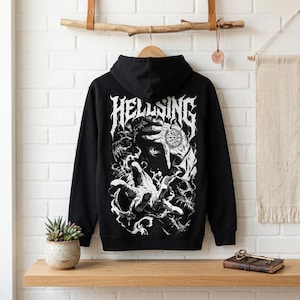 Peut inclure: Sweat à capuche noir avec un motif blanc détaillé d'une figure avec une main tendue, un crâne et le mot "HELLSING" en police gothique. Le sweat à capuche est suspendu à une tringle en bois.