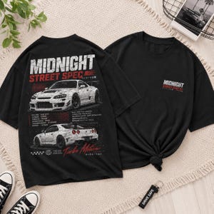 May include: Black t-shirts with a car graphic. One shirt features the text "MIDNIGHT STREET SPEC" with car illustrations and technical details. The other shirt has the text "MIDNIGHT STREET SPEC" on the front. A keychain that says "DRIVE SAFE" is also visible.