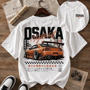 May include: White t-shirt featuring a graphic of an orange sports car with a large spoiler. The shirt has the word "OSAKA" in large black letters, with "Night Shift" in orange. Additional text includes Japanese characters and the words "Built for the streets. Driven by passion."
