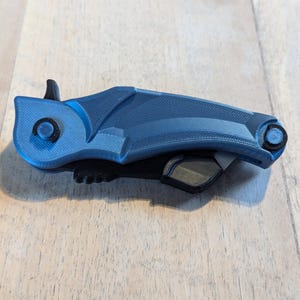 May include: A blue and black 3D-printed object with a unique, angular design. The object has a textured surface and features a circular element on one end. The color scheme is primarily blue with black accents, creating a modern aesthetic.