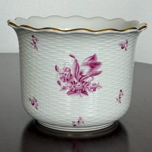 May include: A white porcelain planter featuring a textured basket-weave pattern on the exterior. The vessel is decorated with small magenta floral sprigs and a larger central floral bouquet. The scalloped rim is finished with a thin gold trim.
