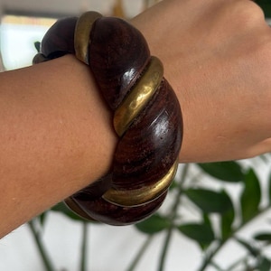 Op de afbeelding: Een dikke armband met een gedraaid ontwerp dat afwisselt tussen segmenten van donkerbruin hout met een zichtbare nerf en glad, gepolijst goudkleurig metaal. De armband heeft een dik, afgerond profiel en een doorlopende cirkelvorm.
