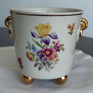 Può includere: Un vaso di ceramica bianca con dettagli dorati, con un motivo floreale con fiori gialli, viola e rosa. Il vaso ha due manici dorati e tre piedini dorati. Una linea nera borda il bordo superiore.