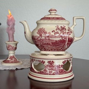 Può includere: Una teiera color crema con un design a scena pastorale rosso, appoggiata su uno scaldavivande abbinato. A sinistra, una candela rosa accesa in un portacandele decorativo. La teiera ha un manico e un coperchio.