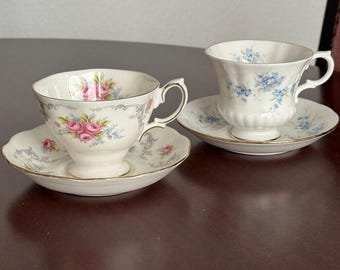 May include: Two porcelain teacups and matching saucers with gold rims. One cup features pink roses and blue flowers, while the other displays blue floral patterns. Both pieces have scalloped edges and delicate handles on a white base.