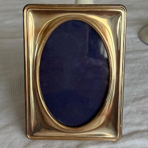 Puede incluir: Un marco de fotos rectangular dorado con una abertura ovalada. El marco tiene un respaldo azul oscuro. El marco está sobre una superficie blanca.