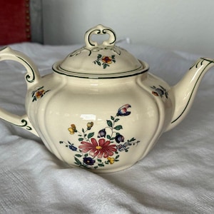 Könnte beinhalten: Eine cremefarbene Keramik-Teekanne mit geriffeltem Körper und passendem Deckel mit dekorativem Griff. Die Teekanne ist mit einem bunten Blumenmuster in Rot-, Blau-, Gelb- und Grüntönen verziert und weist grüne Ränder an den Kanten auf.