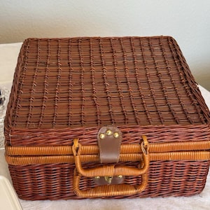 Peut inclure: Un panier de pique-nique rectangulaire en osier tressé marron, doté d'un couvercle à charnière, d'une poignée intégrée robuste et d'une attache centrale en similicuir marron fixée par des rivets métalliques dorés au tissage uniforme.