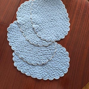 May include: A set of four oval-shaped placemats in a light blue color. Each placemat features a quilted texture with a cross-hatch pattern and a scalloped edge finished with white piping trim. The placemats are arranged in an overlapping stack.