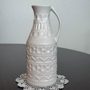 May include: A light gray ceramic pitcher with a handle and textured geometric patterns. The pitcher is placed on a white lace doily. The design includes circles, lines, and zigzags. The pitcher is a decorative item.