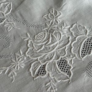 Puede incluir: Un tejido blanco con un intrincado bordado tono sobre tono de rosas en flor y pequeñas flores. El diseño incorpora elementos de calado con patrones de rejilla abiertos y bordes festoneados, mostrando un detallado trabajo de aguja.