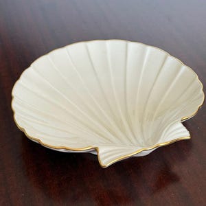 May include: Cream-coloured, shell-shaped dish with a gold trim. The dish features a ribbed design and is set against a dark brown background. A decorative piece for coastal decor.
