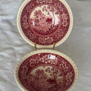 Może przedstawiać: Dwie okrągłe ceramiczne miski z ciemnoczerwonym wzorem transferowym na kremowym tle. Wzór zawiera centralny krajobraz wiejski z drzewami i budynkami, otoczony szeroką bordiurą ze szczegółowymi motywami kwiatowymi oraz koralikowym brzegiem.
