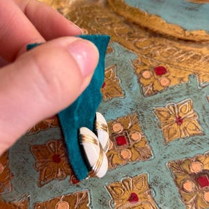 Puede incluir: Un par de pendientes vintage con un centro ovalado blanco y ribete dorado. Los pendientes se sostienen contra una superficie de terciopelo verde azulado, con un fondo decorativo dorado y verde azulado.