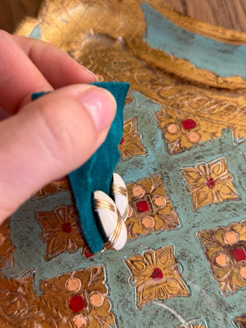 Puede incluir: Un par de pendientes vintage con un centro ovalado blanco y ribete dorado. Los pendientes se sostienen contra una superficie de terciopelo verde azulado, con un fondo decorativo dorado y verde azulado.