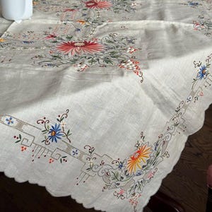 May include: Cream-coloured embroidered tablecloth with a scalloped edge. The floral embroidery features red, orange, blue, and green flowers and leaves. The tablecloth is spread out on a dark wooden table.