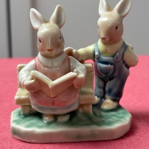 Op de afbeelding: Een keramieken beeldje met twee konijnen. Eén konijn zit op een bank en leest een boek, de ander staat ernaast. Het beeldje heeft pastelkleuren, met konijnen in lichtroze en blauwe overalls.