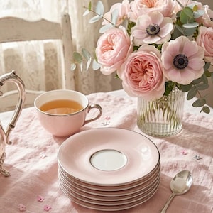 May include: A stack of light pink ceramic saucers with silver rims, a matching light pink teacup filled with tea, a silver spoon with a sugar cube, and a glass vase containing a bouquet of light pink roses and anemones with eucalyptus.