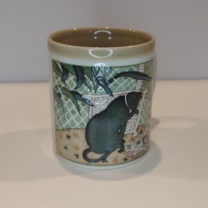 May include: A ceramic mug with a cylindrical shape. The mug features a green and white design with a stylized illustration of a dark green animal. The rim and base of the mug are a light beige color.
