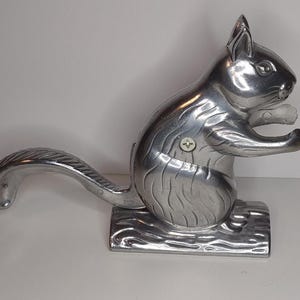 May include: A silver-colored squirrel nutcracker. The squirrel is in a sitting position with its arms outstretched. The tail curves upward. The nutcracker is mounted on a base that resembles a log.