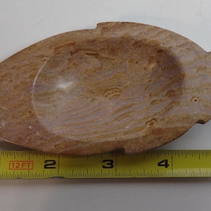 May include: A leaf-shaped stone dish with a polished tan and brown marbled surface. A yellow measuring tape is positioned along the bottom edge, indicating the object is approximately 6 inches in length from tip to tip.