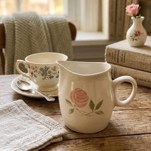 May include: A cream-colored ceramic creamer pitcher featuring a painted pink rose with green leaves, accompanied by a matching teacup on a saucer with a blue floral pattern, a silver spoon, and a small bud vase with a single pink rose.