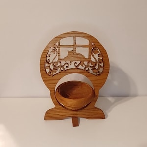 May include: A light brown wooden stand featuring a circular cutout with a carved silhouette of a cat sitting in a window frame, surrounded by decorative scrollwork. A small, matching wooden bowl is positioned within the lower circular opening.