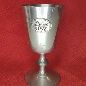 May include: A silver-colored goblet with a wide, rounded bowl and a pedestal base. The goblet has a decorative oval emblem with the letters "OSV". The background is a solid red color.