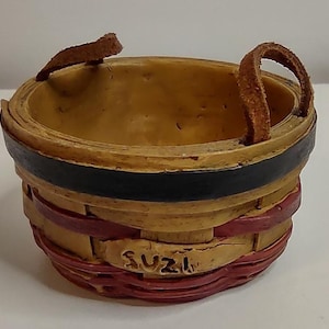 May include: A small round basket with a tan interior and a woven-look exterior featuring a black band at the top and red accents at the base. The basket has two brown leather-like handles and a small plaque on the front with the name SUZI inscribed.