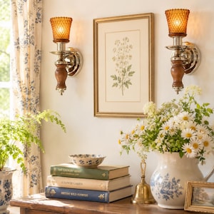 May include: Two wall-mounted sconces with amber-colored textured glass shades, silver metal bases, and wooden accents. Between them hangs a framed botanical print. Below, a stack of three books, a small bowl, a brass bell, and a floral pitcher.