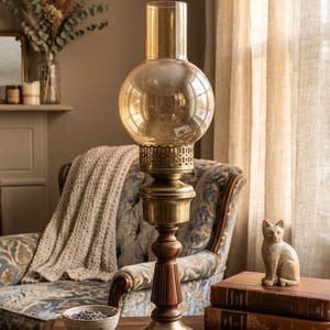 May include: A vintage-style oil lamp featuring a round amber-tinted glass chimney, a decorative brass collar with cut-out details, a central brass section, and a dark wooden pedestal base that sits on a flat, circular brass foundation.