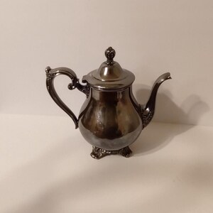 May include: A polished dark metallic teapot featuring a rounded body, a curved handle with ornate detailing, a slender spout, and a lid topped with a small finial. The teapot stands on three decorative feet and has a reflective, smooth surface.