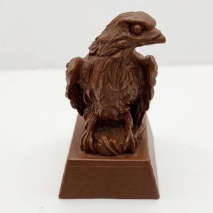 May include: A brown molded plastic figurine of an eagle perched on a rock, set upon a rectangular base. The eagle is depicted with detailed feathers, a sharp beak, and focused eyes, captured in a solid, monochromatic brown finish throughout.