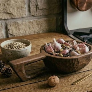 May include: A dark brown carved wooden bowl with a handle containing garlic cloves and green cardamom pods, accompanied by a small speckled beige ceramic bowl filled with coriander seeds, a single walnut, and a gold-toned metal spoon.