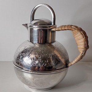 May include: A silver-colored, vintage-style pitcher with a spherical body and a woven handle. The pitcher features an ornate floral pattern etched into the metal. It has a lid and a curved spout for pouring.