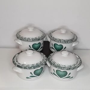 May include: A set of four white ceramic lidded pots, each featuring a green heart design on the front and a decorative green and white speckled pattern along the rim of the lid. Each pot includes two side handles and a small knob on the lid.