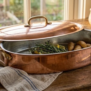 May include: An oval copper roasting pan with a silver-toned interior, two brass side handles, and a matching copper lid with a brass handle. The pan contains fresh rosemary sprigs and small potatoes, resting on a light-colored linen cloth.