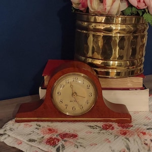 May include: A wooden mantel clock with a curved top and a gold-toned circular face featuring black Arabic numerals and the text Jack Thomas. The clock has a polished finish and a thin gold-colored trim along the base, displaying a classic design.