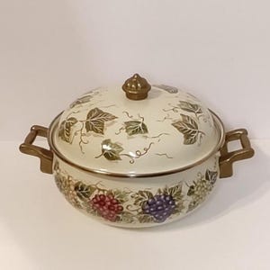 May include: A round, cream-colored enameled metal casserole dish with a matching lid. The pot features a decorative pattern of green grape leaves and clusters of red and purple grapes. It has two gold-toned side handles and a gold-toned lid knob.